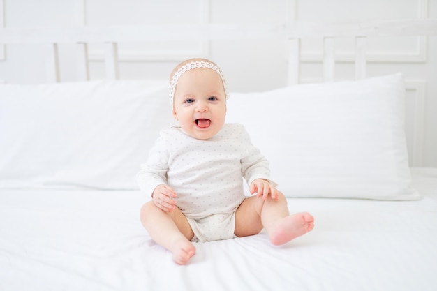 Een babymeisje van zes maanden zit thuis op een wit katoenen bed in een bodysuit op het bed en lacht