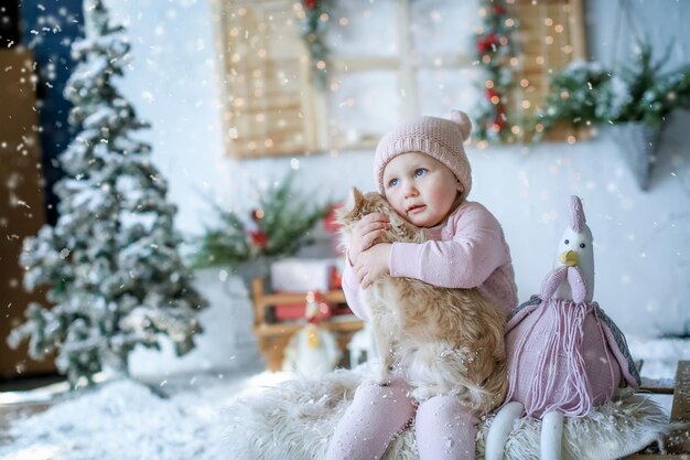 Een babymeisje zit in een studio met kunstmatig winterlandschap in een roze trui en een roze muts.