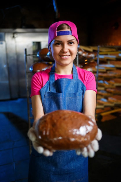 Een bakkersmeisje neemt warm brood in een bakkerij tegen de achtergrond van planken met brood. Industriële productie van brood. Het stadium van bakken in een bakkerij