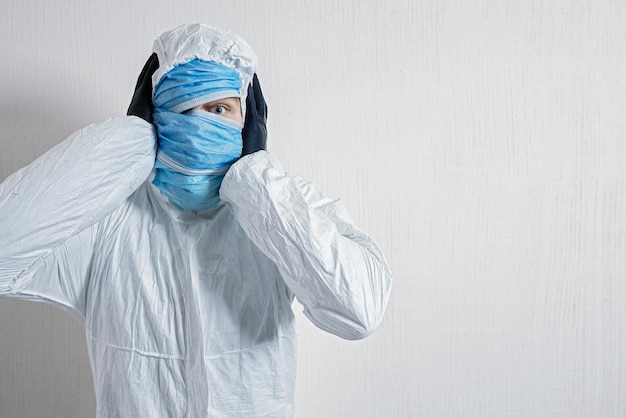 Een bange man in een beschermend pak hing met medische maskers tegen een witte muur. De verschrikkingen van de epidemie, het gevaar van het coronavirus. de wetenschapper houdt zijn hoofd vol afschuw