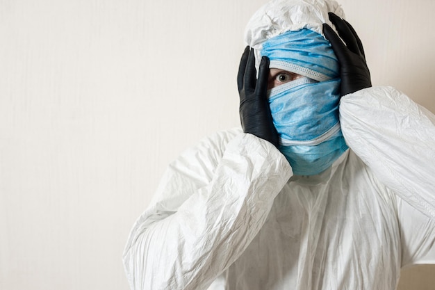 Een bange man in een beschermend pak met medische maskers beeldt afschuw af tegen een witte muur. de verschrikkingen van de epidemie, het gevaar van het coronavirus