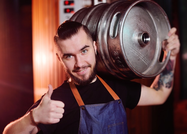 Een bebaarde brouwer in een schort houdt een metalen vat met bier in zijn handen en glimlacht