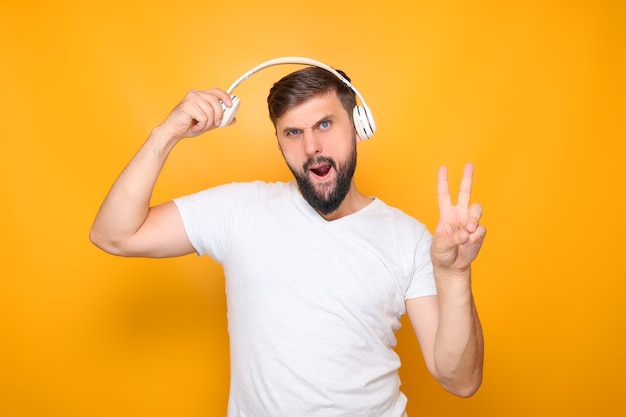 Een bebaarde man in een wit t-shirt, die op een gele achtergrond staat, luistert naar muziek op een koptelefoon.