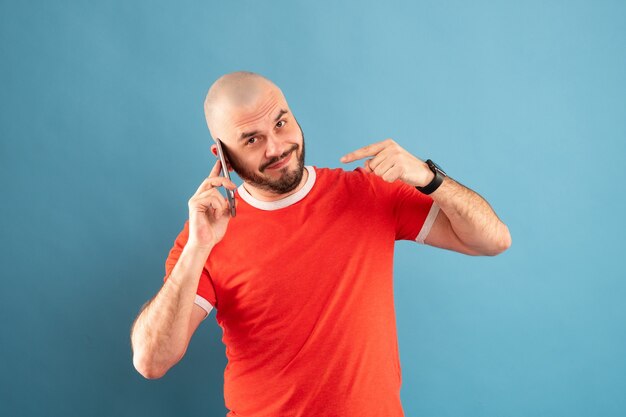 Een bebaarde man van middelbare leeftijd in een rood T-shirt op een blauwe achtergrond wijst met zijn hand naar de telefoon. Bel me. Geïsoleerd.