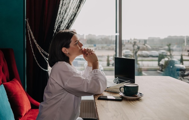 Een bedachtzame brunette zakenvrouw in een witte blouse zit aan een tafel in een café met een telefoon en laptop