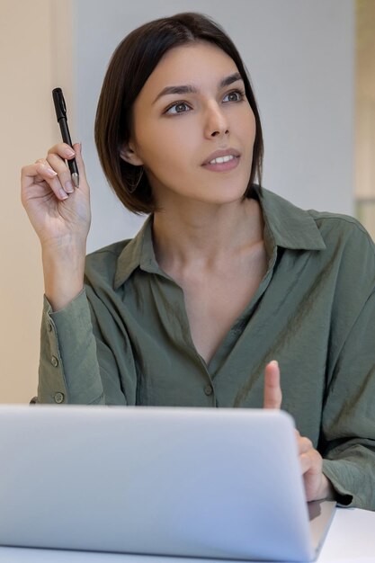 Een bedachtzame jonge vrouwelijke bedrijfsmedewerker met een pen in de hand die bij de computermonitor zit
