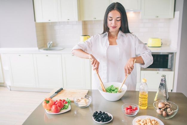 Een beeld van brunette die ingrediënten in witte kom mengt met twee houten lepels. ze staat in de keuken. er zijn veel ingrediënten aan tafel.