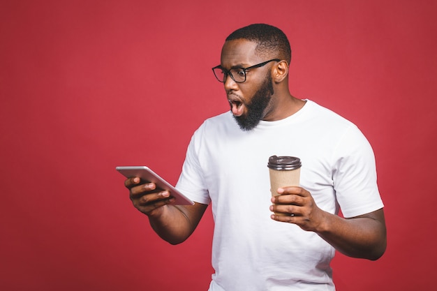 Een bericht typen. Verraste zwarte mens die iets op de mobiele telefoon typen, die koffie drinken terwijl status geïsoleerd tegen rode achtergrond.