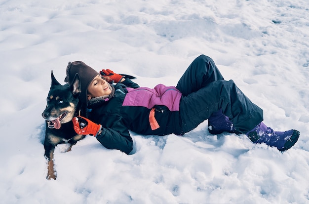 Een blank meisje dat erg warm was en een hoed draagt die met haar hond in de sneeuw ligt.