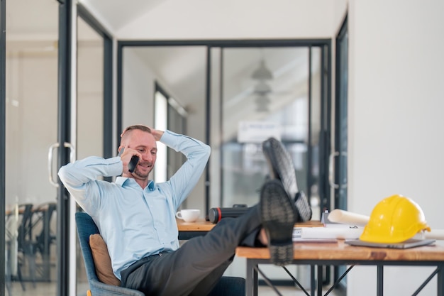 Foto een blanke ingenieur die zijn benen op de tafel legt en op de mobiele telefoon praat met klanten in het kantoor.