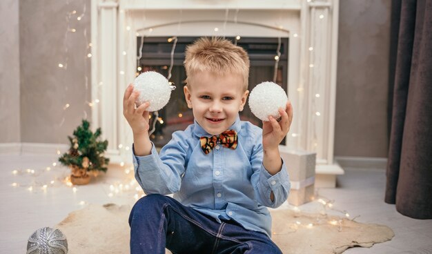 Een blanke kleine jongen in een blauw shirt en een vlinderdas van een tijger zit bij de open haard in een kamer met kerstballen