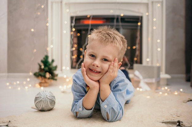 Een blanke kleine jongen in een blauw shirt met een vlinderdas ligt op een pelskleed bij de nieuwjaarsopen haard