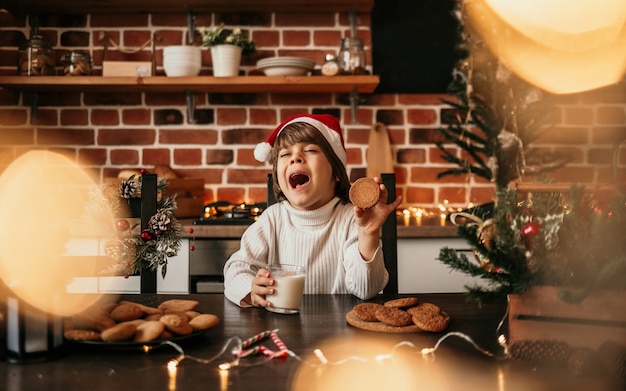 Een blanke kleine jongen in een witte gebreide trui en een rode kerstmuts zit aan de keukentafel met havermoutkoekjes en melk