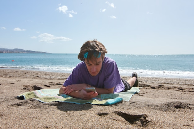 Een blanke man liggend op een handdoek op een strand die overdag zijn telefoon checkt