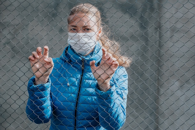 Een blanke meisje in een beschermend medisch masker staat achter een metalen hek. Strikte woningisolatie tijdens quarantaine.