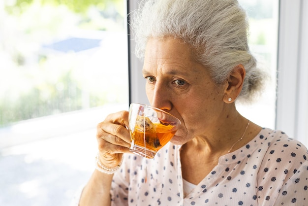 Een blanke oudere vrouw drinkt thee in het raam van een zonnig huis.