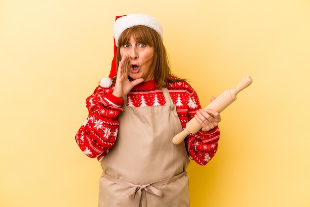 Een blanke vrouw van middelbare leeftijd die koekjes kookt voor Kerstmis geïsoleerd op een gele achtergrond, zegt een geheim heet remnieuws en kijkt opzij