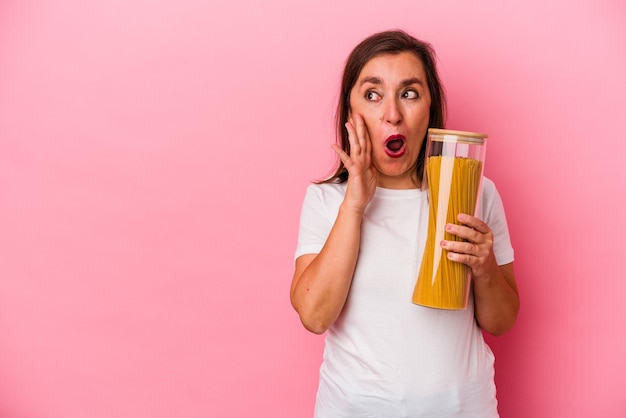 Een blanke vrouw van middelbare leeftijd met een pastapot geïsoleerd op een roze achtergrond, zegt een geheim heet remnieuws en kijkt opzij looking