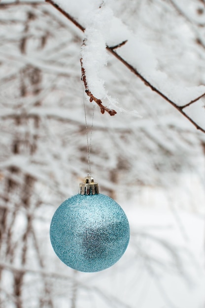 Een blauwe glanzende bal hangt aan een besneeuwde tak op een wazige achtergrond in een verticaal formaat