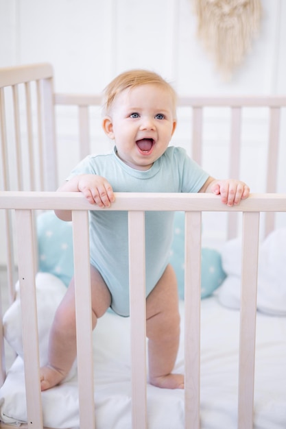 Een blije lachende babyjongen staat thuis in een wieg in blauwe kleren een klein kind leert staan of wordt 's ochtends wakker een gelukkige jeugd en gezin