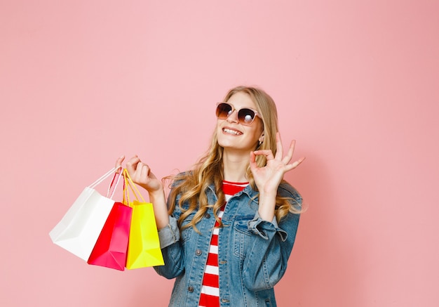 Een blond meisje dat blij is met het winkelen dat ze heeft gedaan en gebaar over roze achtergrond toont