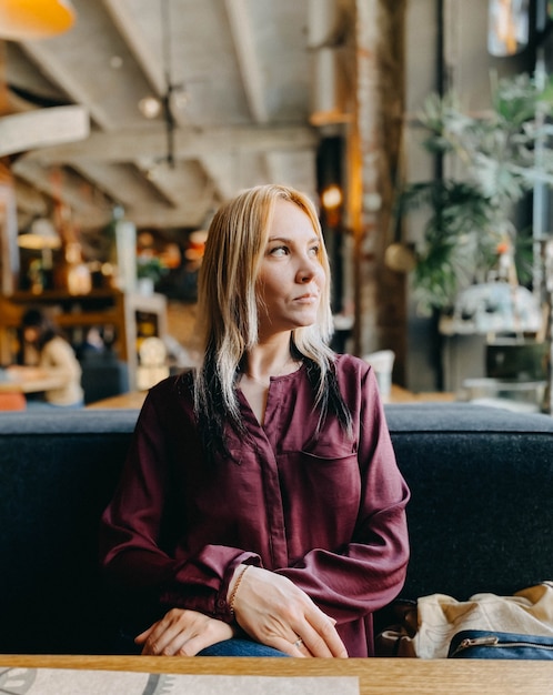 Een blond meisje in een lila trui zit in een gezellig restaurant op een bank en ze is ontspannen en glimlachend.