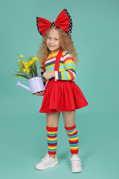 een blond meisje in een rok, kniekousen en een T-shirt met een rode strik houdt een emmer bloemen vast