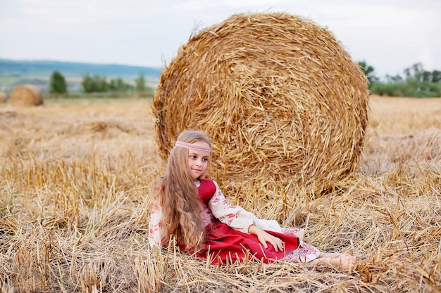 een blond meisje met grote bruine ogen in een Slavisch kostuum zit op een veld bij een hooiberg