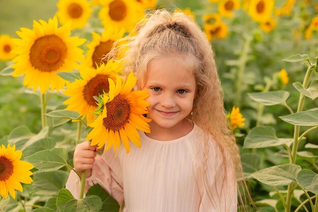 een blond meisje met lang haar in een roze linnen jurk staat in een veld met zonnebloemen