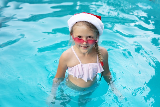 een blond meisje staat in het zwembad in een roze bril met kerstmanhoed en lacht nieuwjaar