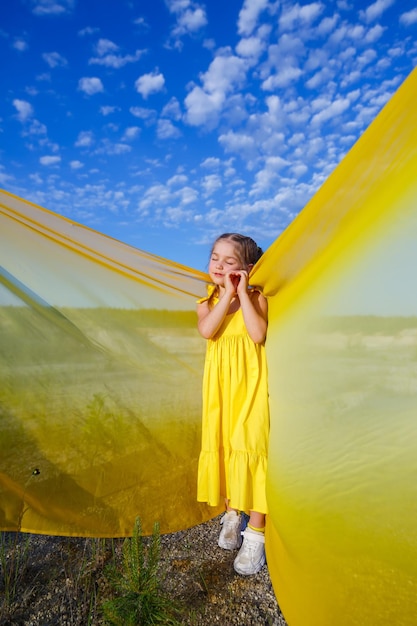 Een blond meisje van 7-8 jaar oud in een felgele jurk tegen een blauwe lucht.