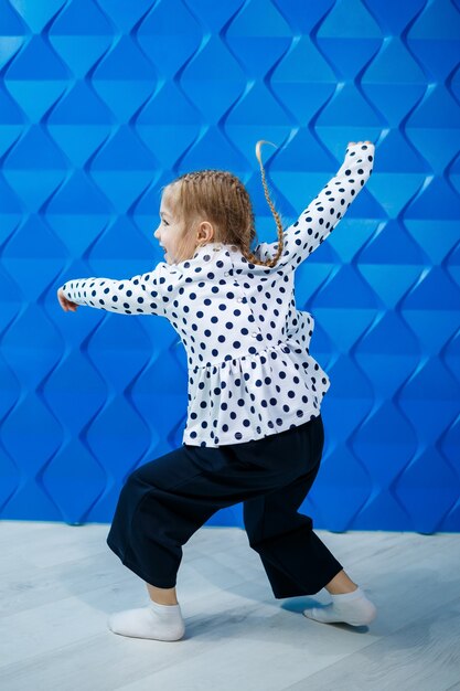 Een blond meisje van 7 jaar oud in een jasje met erwten op een blauwe achtergrond danst en lacht, heldere kinderemoties van vreugde, een gelukkige jeugd