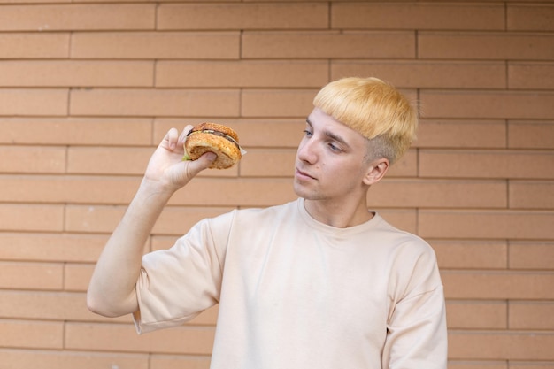 Een blonde Europese man gekleed in een beige T-shirt met een hamburger in zijn hand en ernaar kijkt