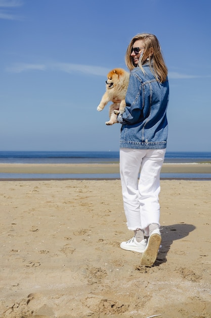Een blonde jonge vrouw in een jeansjasje en witte broek die kleine pomeranian hond bij een zandstrand houdt