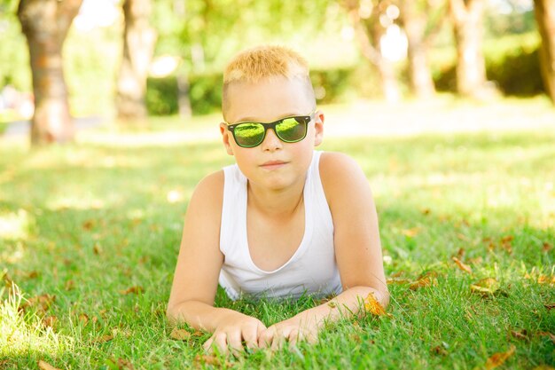 Een blonde jongen in een wit T-shirt met zonnebril ligt op het gras in het park