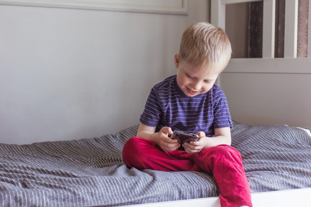 Een blonde jongen zit op het bed en houdt de smartphone in zijn handen Online communicatie videocommunicatie onderwijs