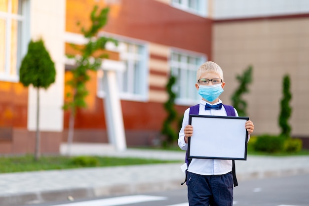 Een blonde schooljongen met bril en een beschermend masker staat op de school en houdt een bord vast met een wit laken.