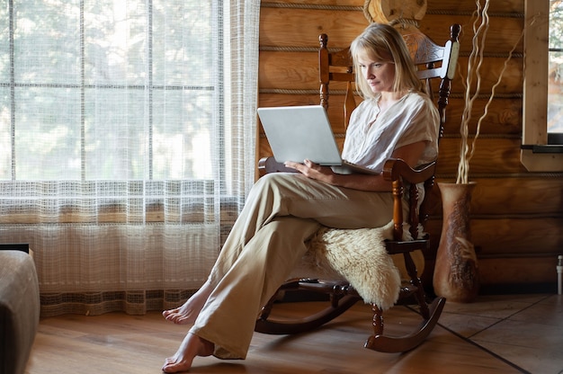 Een blonde vrouw in beige huiskleren zit in een schommelstoel met een laptop in haar handen in een gezellige ro...