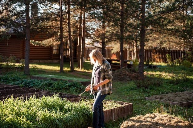 Foto een blonde vrouw in een geruit overhemd met handschoenen en een hark in haar handen staat in de achtertuin in de zon...