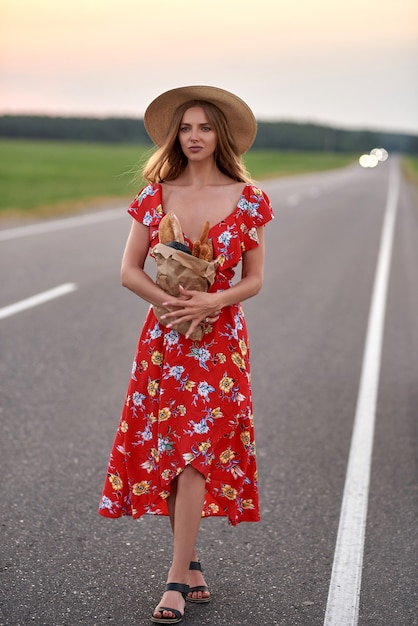 Een blonde vrouw in een rode jurk en een strohoed staat langs de weg