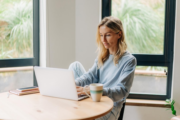 Een blonde vrouw in huiskleding zit thuis aan een tafel en werkt hard aan een laptop op een externe baan