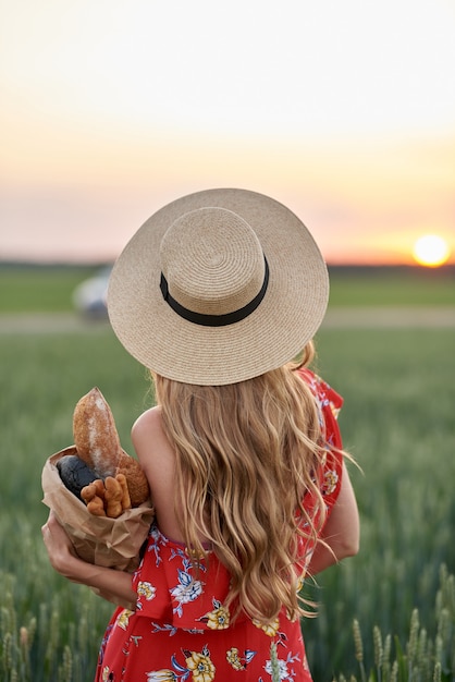 Een blondine in een rode jurk en een strohoed met stokbrood in haar handen op een tarweveld bij zonsondergang