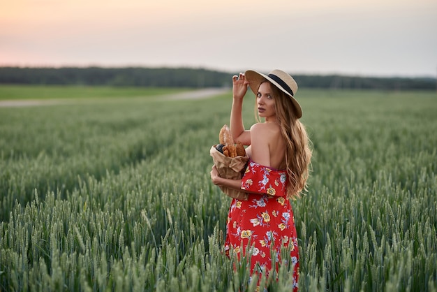 Een blondine in een rode jurk en een strohoed met stokbrood in haar handen op een tarweveld bij zonsondergang