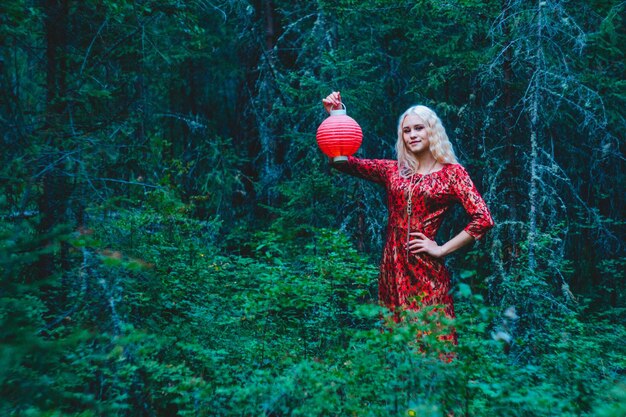 Een blondine in een rode jurk in het bos met een rode Chinese lantaarn in haar handen