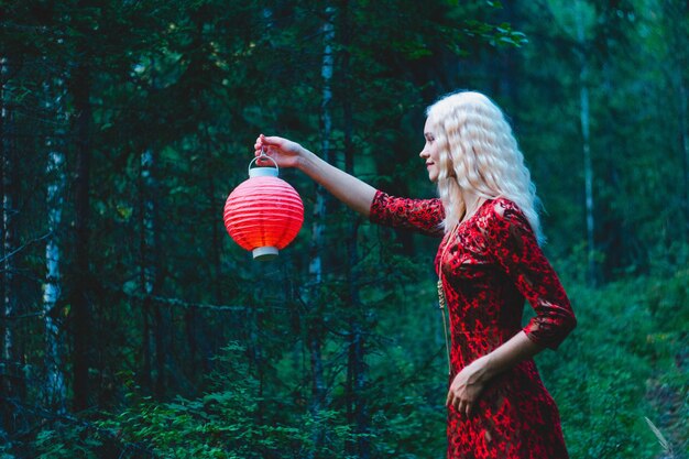 Een blondine in een rode jurk in het bos met een rode Chinese lantaarn in haar handen