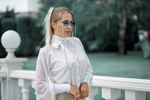 Een blondine in een witte blouse en zonnebril poseert tegen de achtergrond van een groene tuin