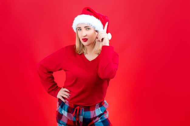 Een blondine met een kerstmuts van de kerstman en een nieuwjaarspyjama houdt haar wijsvinger bij haar gezicht