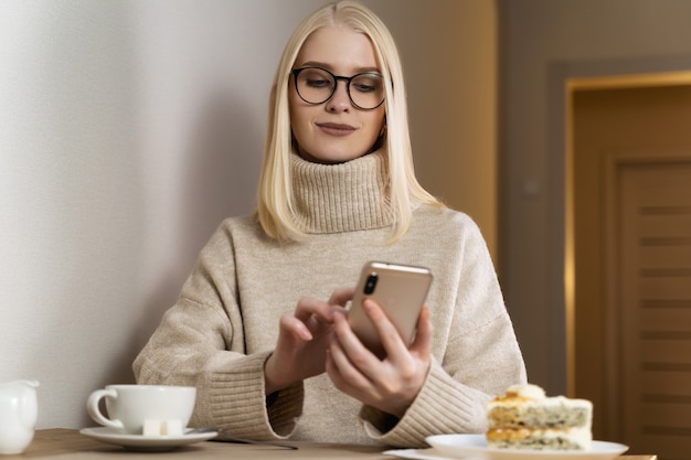 Een blondine met los haar en een bril met een zwart montuur zit aan een tafel in een warme beige trui.