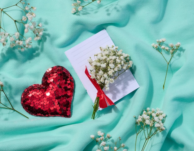 Een boeket van gypsophilia een rood hart gemaakt van pailletten en een gevouwen wit vel papier op een achtergrond van textiel een notitie Bekijk van bovenaf