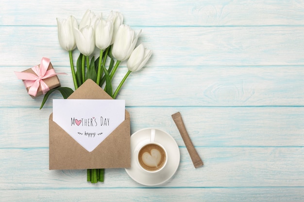 Een boeket van witte tulpen, kopje koffie, geschenkdoos met een liefdesbrief en envelop op blauwe houten planken. Moederdag